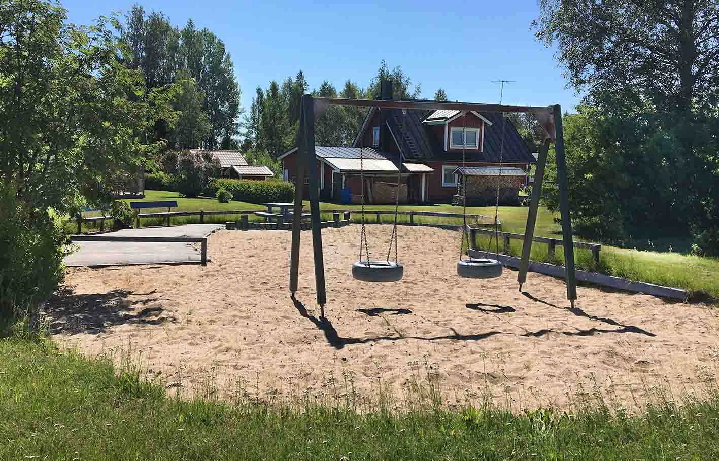 Två gungor i en gungställning med sand under. Längre bak syn sittplatser och bord.