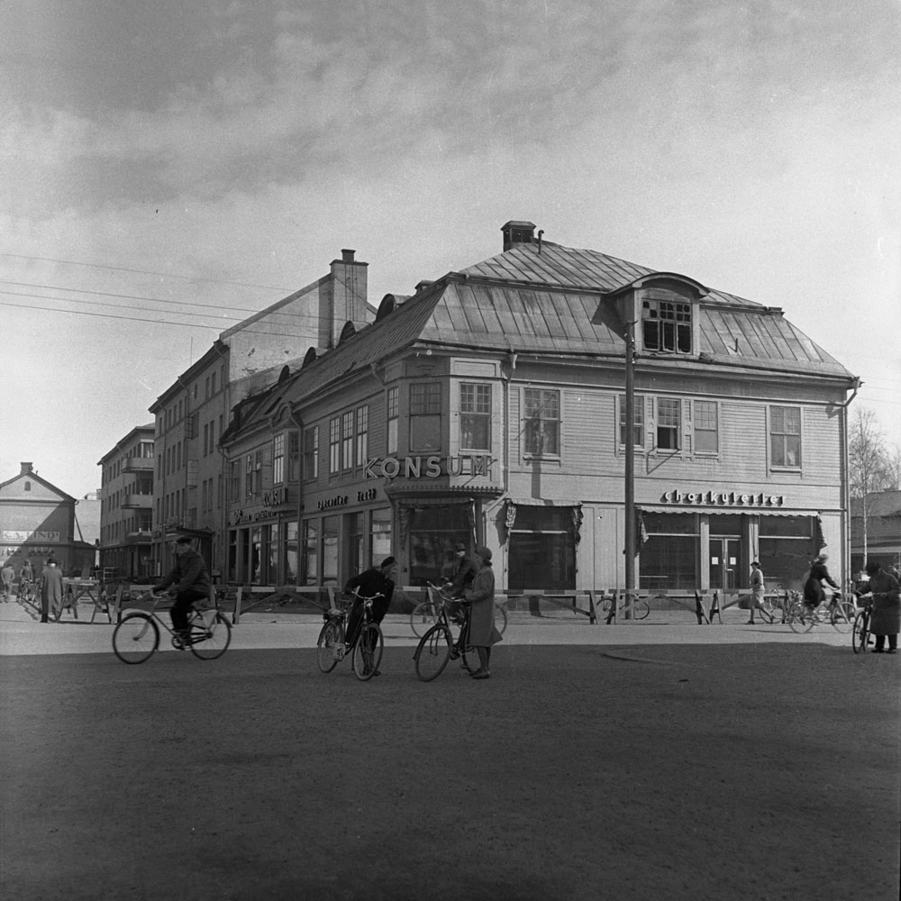 Bild av trähus i gatukorsning, några människor med cyklar framför.