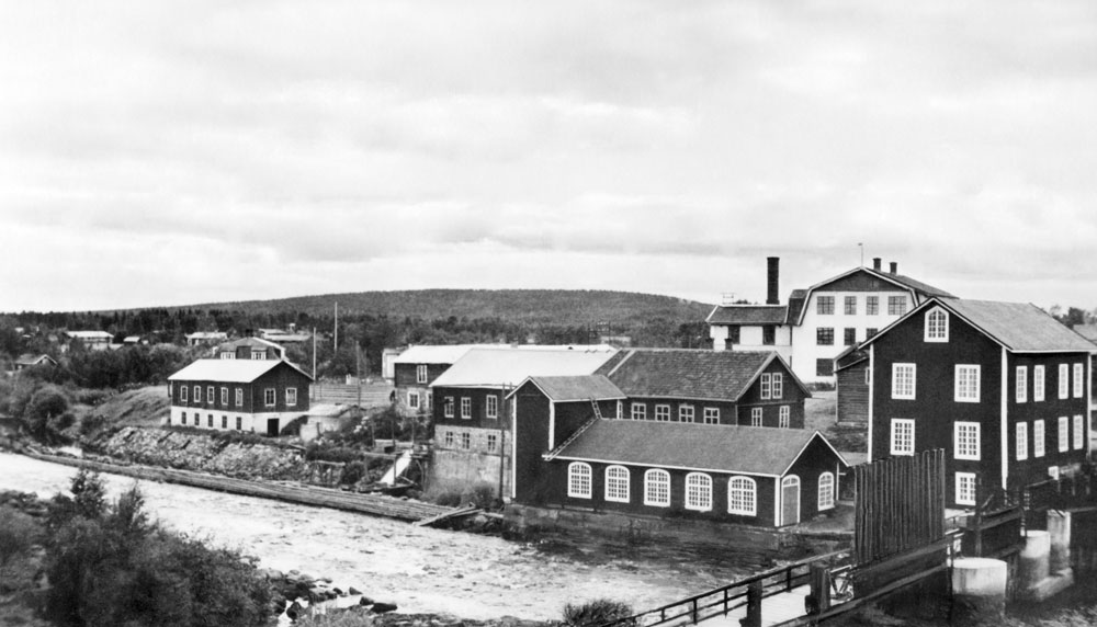 Vy över fabriksområde vid älv, stora träbyggnader med stora fönster.