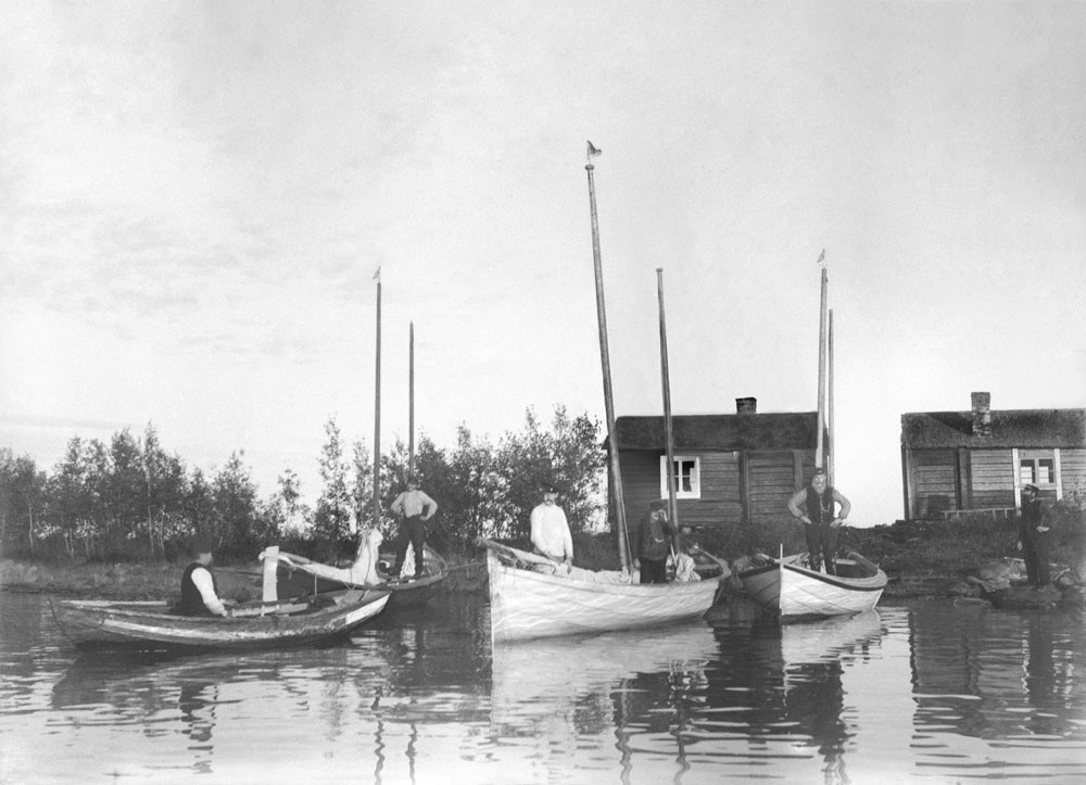 Män i små båtar vid skärgårdsö med två små stugor i bakgrunden.