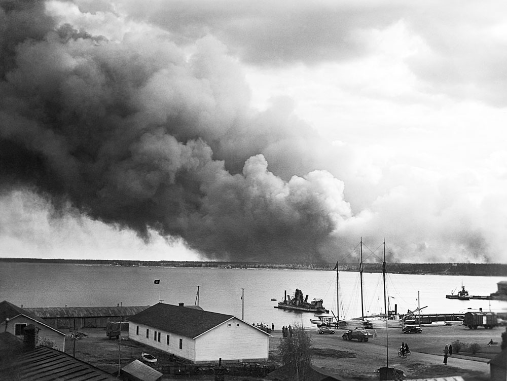 Vy över hamn. I fjärran väller svart rök upp mot himlen.