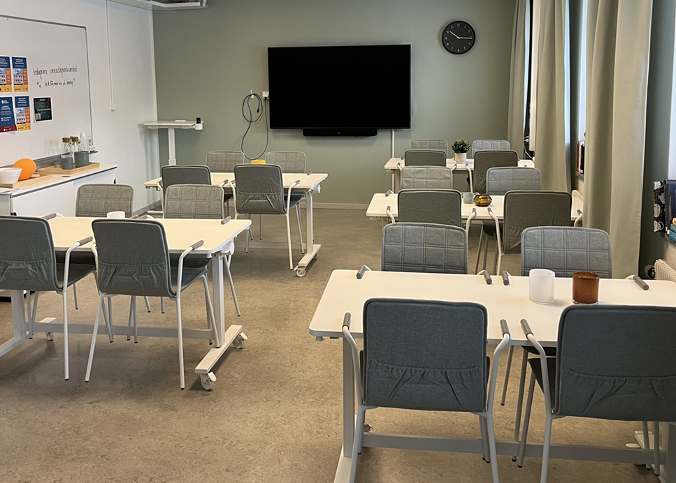 Rum med flera bord och stolar i rader samt stor skärm på väggen.