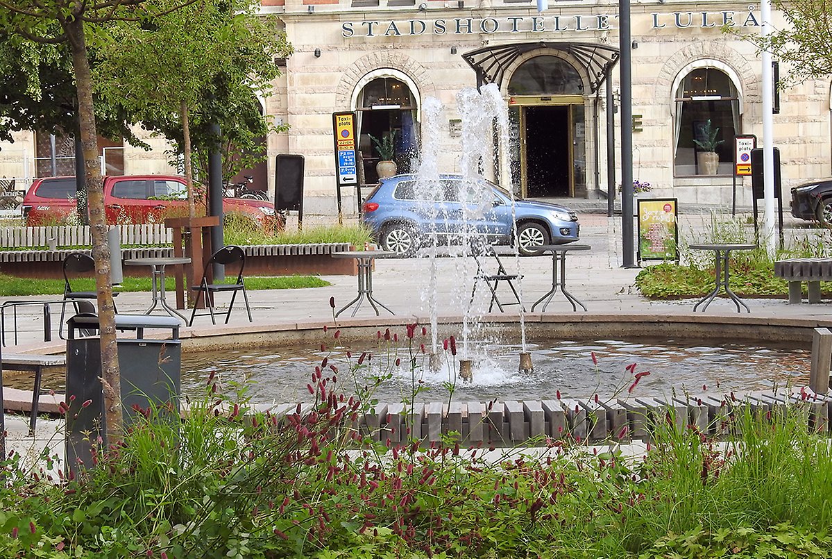 Fontän med stolar och sittplatser runt om. Stadshotellet i bakgrunden och blommor framför